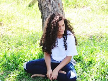 Girl with curly hair looking away while sitting on grassy field by tree in park