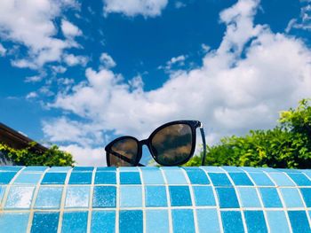 Black sunglasses on the edge of the pool on a day with blue sky and fluffy clouds