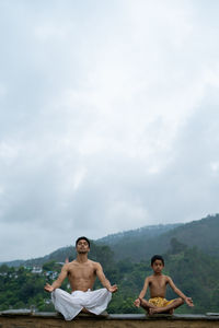 Full length of young man with boy meditating against sky