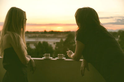 Young woman drinking coffee while sitting on table against sky during sunset