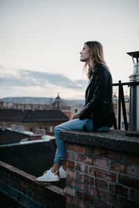 Side view of young woman looking away while sitting on railing against sky