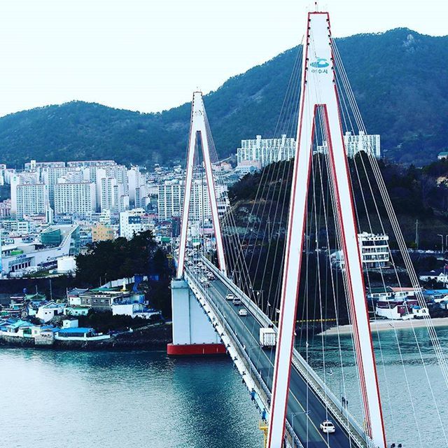 architecture, built structure, transportation, bridge - man made structure, connection, water, river, engineering, mountain, famous place, suspension bridge, nautical vessel, building exterior, travel destinations, waterfront, mode of transport, international landmark, travel, bridge, clear sky