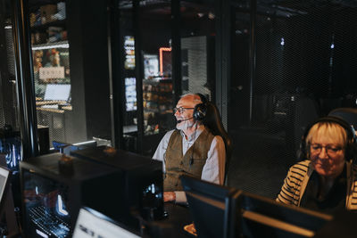 Elderly man wearing headset sitting by female friend playing video game on computer at gaming lounge
