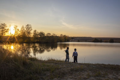 Men standing on lake against sky during sunset