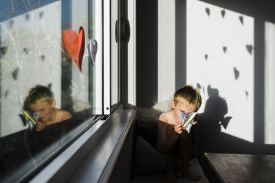 Shirtless boy making decorations while sitting by window at home