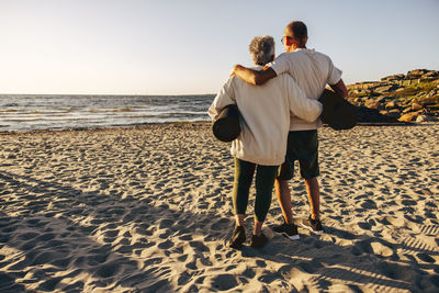 Rear view of senior couple holding exercise mats while standing on sand at beach