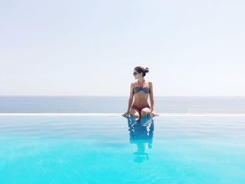 Beautiful woman wearing bikini while sitting amidst swimming pool and sea against clear sky