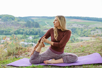Young woman stretching muscles in a park on a yoga mat with a village on a background