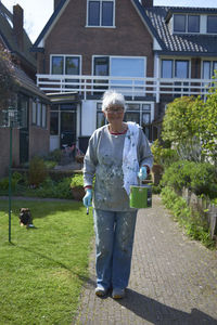 Portrait of senior woman with paint can and paintbrush standing against house
