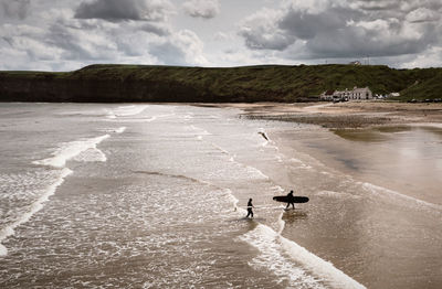 Two surfers walk out of the sea on to an empty beach under a dramatic sky.