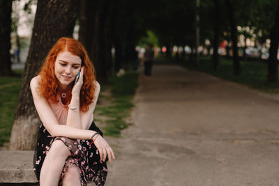 Young redhead woman talking on phone while sitting in park