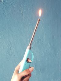 Cropped hand of person holding lit gas lighter against blue wall