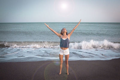 Full length of woman standing with arms raised at beach against sky