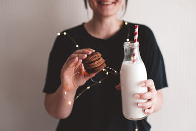 Smiling woman eating chocolate cookies and holding bottle of milk in room close up. winter holiday