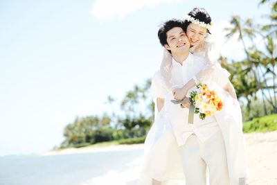 Happy newlywed couple standing at beach against sky during sunny day