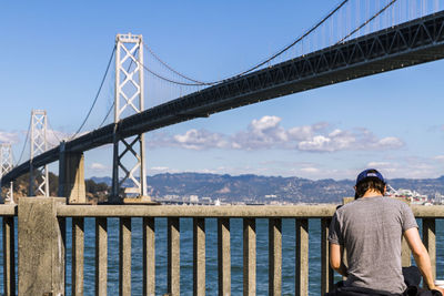 Rear view of man standing by bay bridge over san francisco bay against sky