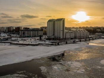 Frozen river by buildings against sky during sunset in city