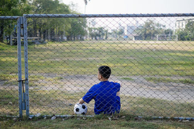 Rear view of teenage boy with soccer ball sitting against chainlink fence