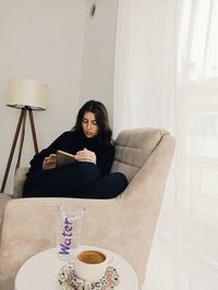 Young woman reading book while sitting on armchair at home