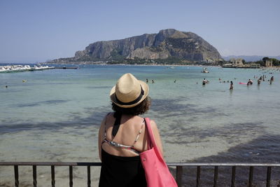 Rear view of woman standing by railing and looking at sea against clear sky during sunny day