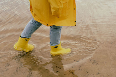 Cheerful young woman in a yellow raincoat and yellow boots steps into the lake with her foot. person