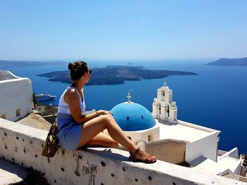 Woman sitting by church and sea against blue sky