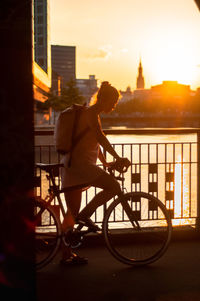 Woman riding bicycle on bridge in city during sunset