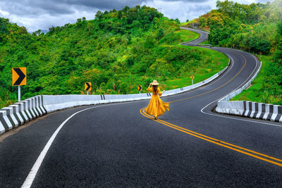 Woman in yellow dress walking on road amidst trees against sky