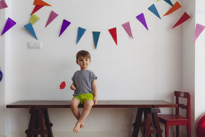 Portrait of cute boy sitting on table against wall at home