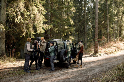 Multiracial male and female friends talking to each other while standing near off-road vehicle on dirt road against tree