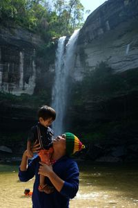 Man carrying son on shoulder while standing against waterfall