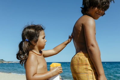 Cute girl applying suntan lotion on brother's back while enjoying sunny day at beach during summer
