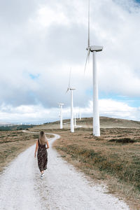 Rear view of woman walking on field by windmills against sky