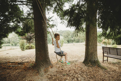 Side view of boy on swing under tree against trees