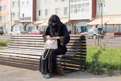 A muslim woman in national clothes sitting on a bench is looking for something  her women's handbag.