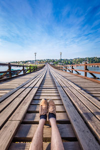 Low section of person sitting on footbridge against blue sky during sunny day