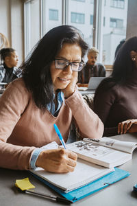 Mature female student writing in book while sitting with friends at classroom