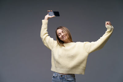 Young woman in yellow sweater on gray background dancing with phone in her hand