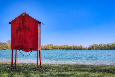 Red built structure by lake against clear blue sky