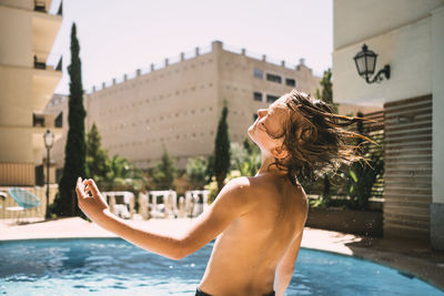 Rear view of shirtless boy enjoying by resort swimming pool against sky