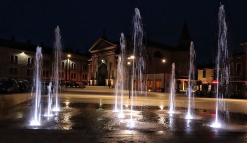 Illuminated fountain by building against sky at night