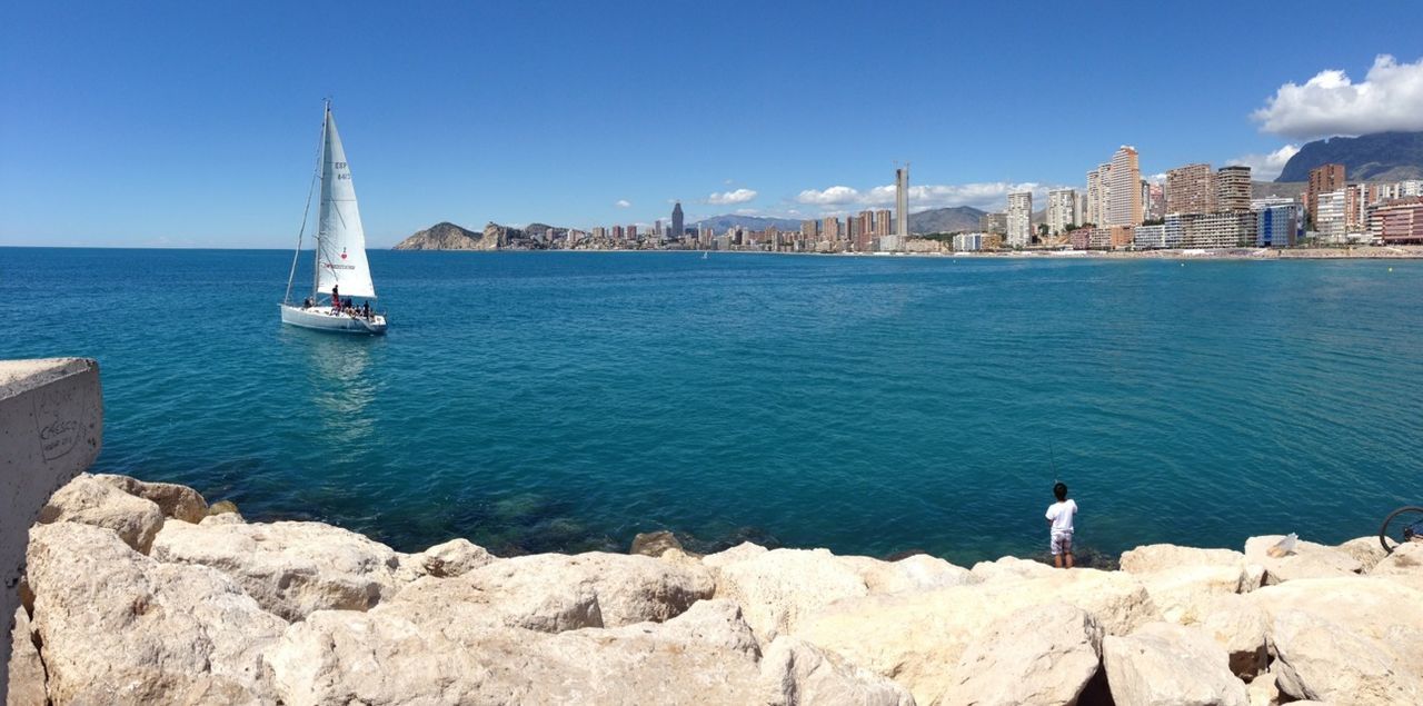 water, sea, nautical vessel, transportation, mode of transport, blue, building exterior, built structure, architecture, boat, men, lifestyles, travel, leisure activity, clear sky, rock - object, rear view, day