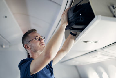 Young man travel by airplane. passenger putting hand baggage in lockers above seats of plane.