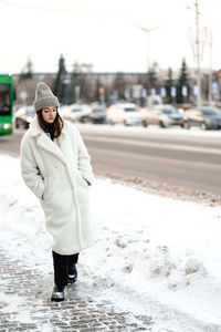 Girl in a fur coat walks along the road traffic. it's cold. the girl has a knitted hat on her head