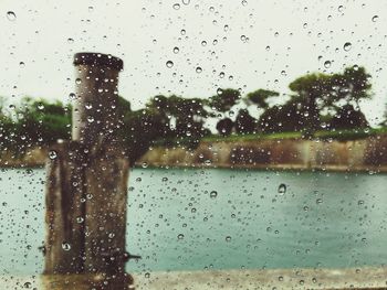 Wooden post in river against sky seen through wet glass window during monsoon