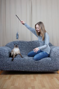 Young woman playing with siamese cat on sofa against wall