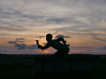 Playful man posing on bench against sky during sunset