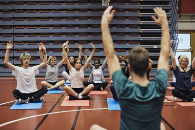 Rear view of male coach with arms raised teaching yoga to students in sports court