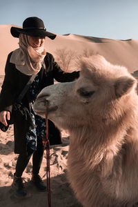 Full length of woman wearing hat on sand dunes getting along with a camel against blue sky