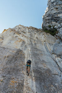 From below back view of unrecognizable mountaineer with safety harness climbing rough steep rocky mountain slope with metal steps